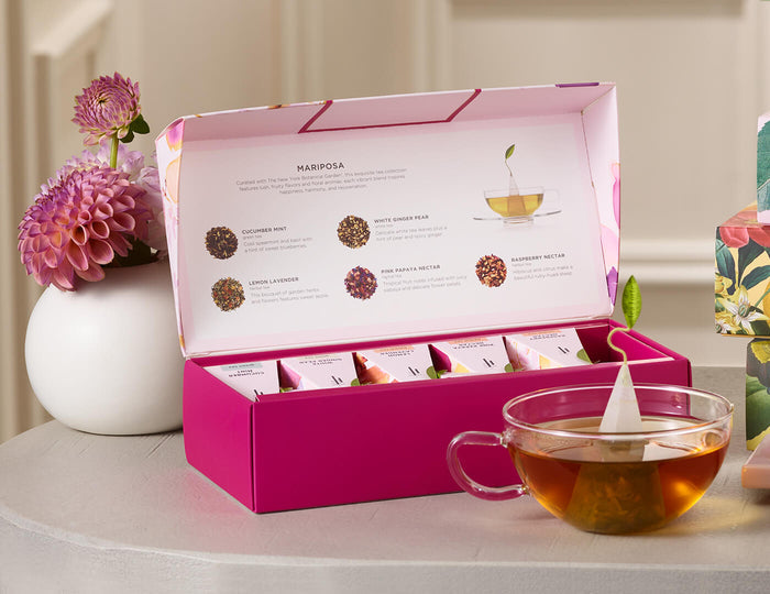 MAIN PDP | Mariposa Petite Presentation Box of 10 pyramid tea infusers, open on a table with a glass cup of tea