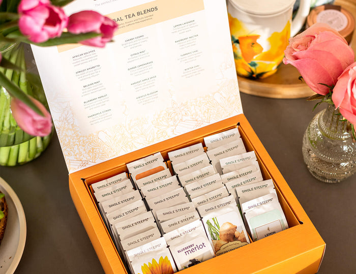 28 count tea chest of Herbal Single Steeps pouches, lid open, showing loose tea pouches inside,  on a table with fresh flowers and a teacup. Photo by @foodie_renee.