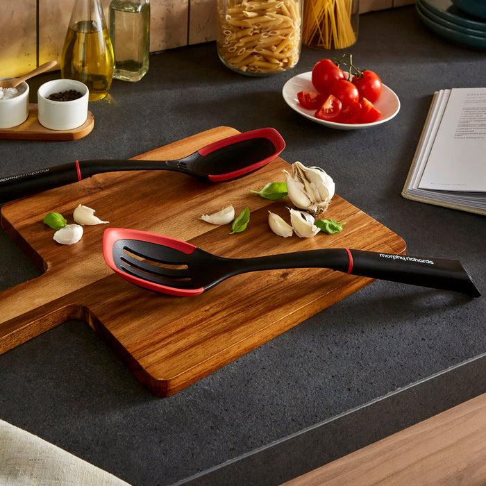 Black and red slotted spoon on a wooden cutting board with garlic and herbs, on a kitchen counter.