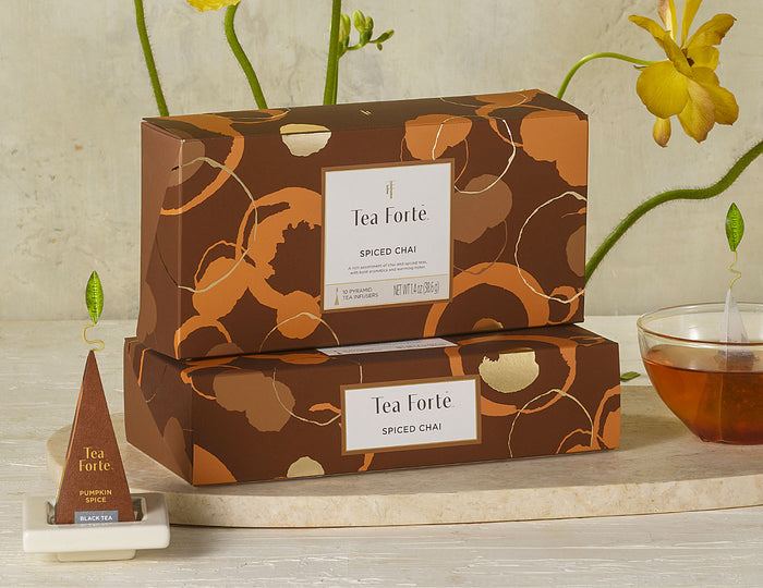 MAIN PDP | 2 Spiced Chai tea assortment boxes in 10 count petites with lid closed, infuser and cup of steeped tea to the side with yellow flowers