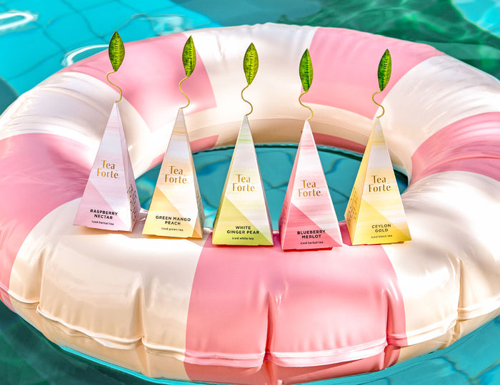 MAIN PDP | Five pyramid-shaped iced teas, each with a green leaf-shaped top, arranged on a pink and white striped inflatable pool float over turquoise water. The tea bags feature different flavor labels including Raspberry Nectar, Green Mango Peach, White Ginger Pear, Blueberry Merlot, and Ceylon Gold.