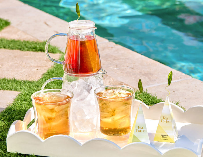MAIN PDP |  Tea Over Ice® Pitcher Set filled with iced tea and two matching POOM glasses with ice, arranged on a white tray beside a pool. Two pyramid tea infuser are visible on the tray.