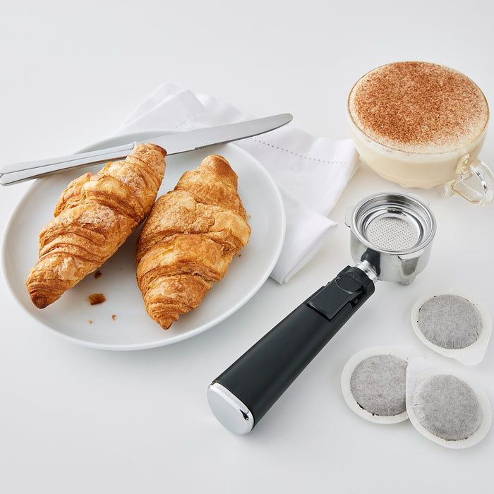 Two croissants on a plate with a coffee cup, press, and filters on a white surface.