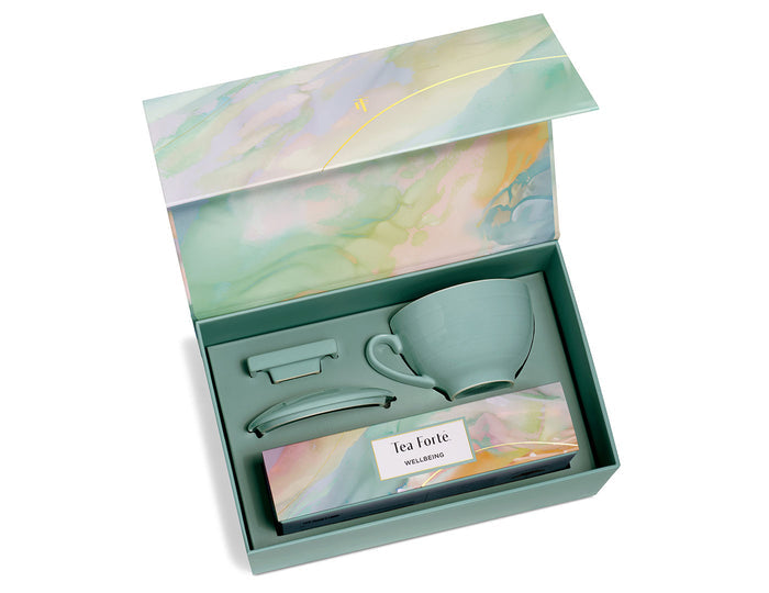 Open gift box containing a gray ceramic mug, tea infuser, spoon, and Tea Forte Wellbeing tea package with watercolor design.